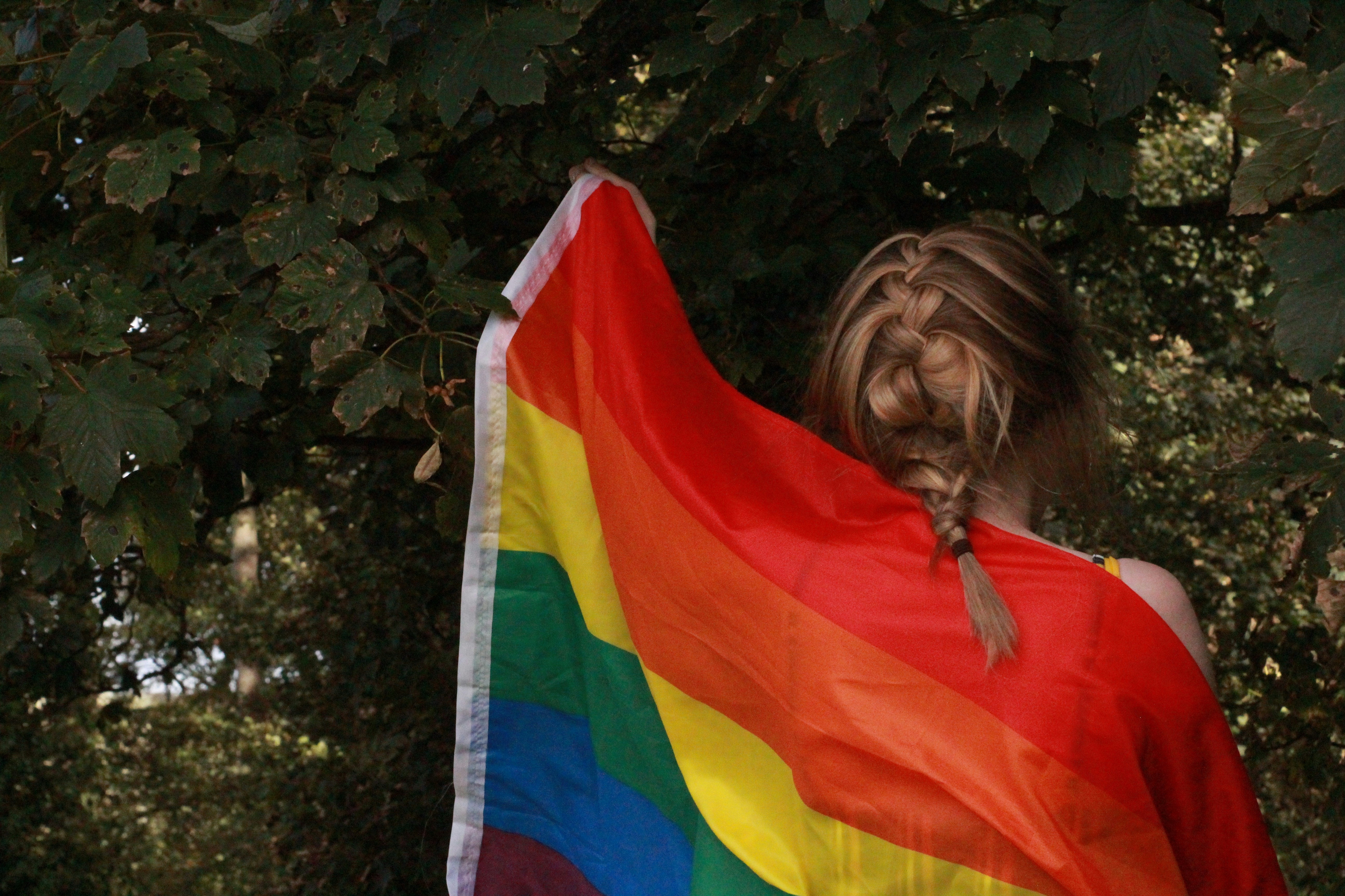 Person with braided hair draped in a rainbow Pride flag, standing in nature