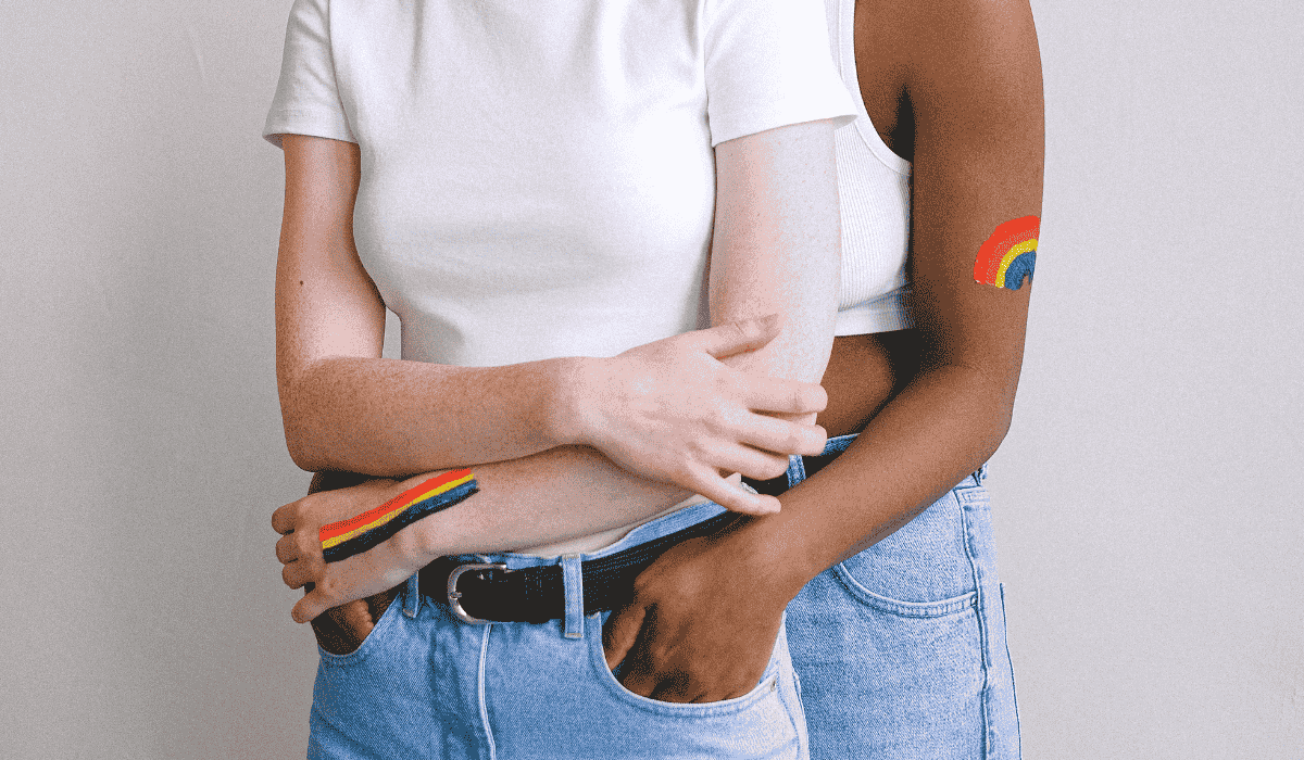 Two women embracing, both wearing pride rainbow body paint and casual white tops and jeans, showing queer love and unity.